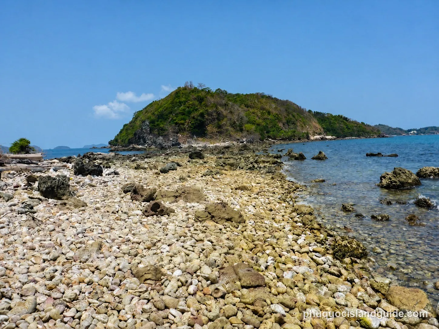 A rocky beach with clear blue water leading to a lush green island under a bright blue sky, with some small clouds.