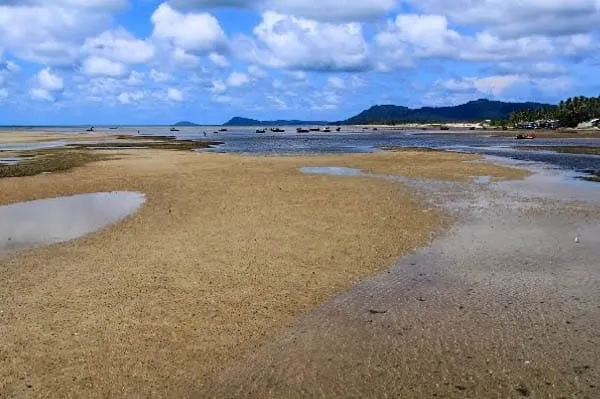 A wide shot of a sandy beach at low tide with shallow water, distant boats, hills, and palm trees on the horizon under a cloudy blue sky.