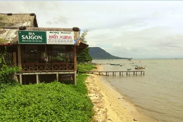 A restaurant with a thatched roof on stilts next to a sandy beach with a wooden pier and boats in the water. A mountain is in the distance under an overcast sky.