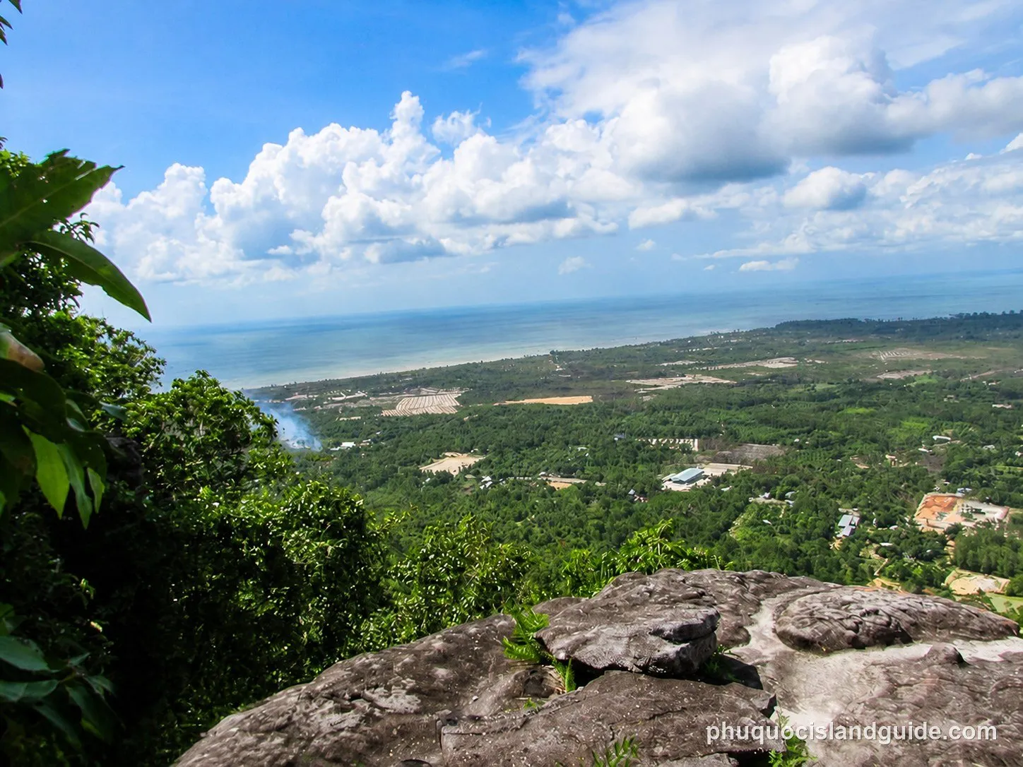 A panoramic view from a rocky outcrop overlooking a lush green tropical landscape, with a coastline and the ocean under a cloudy sky. Smoke rises from the distant forest.