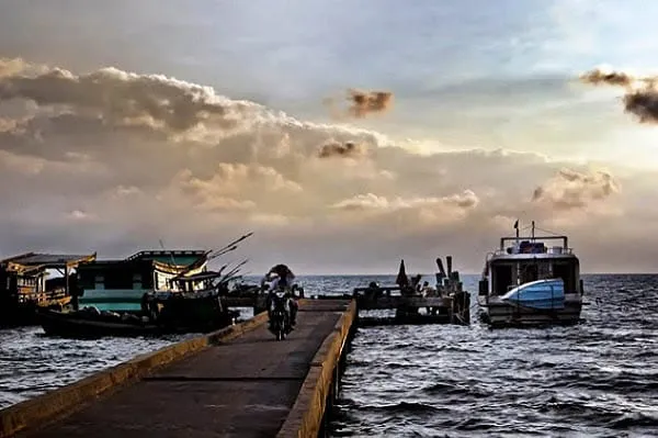 A concrete pier extending into the sea with a person on a motorbike. Several boats and ships are docked on both sides of the pier under a cloudy sky.