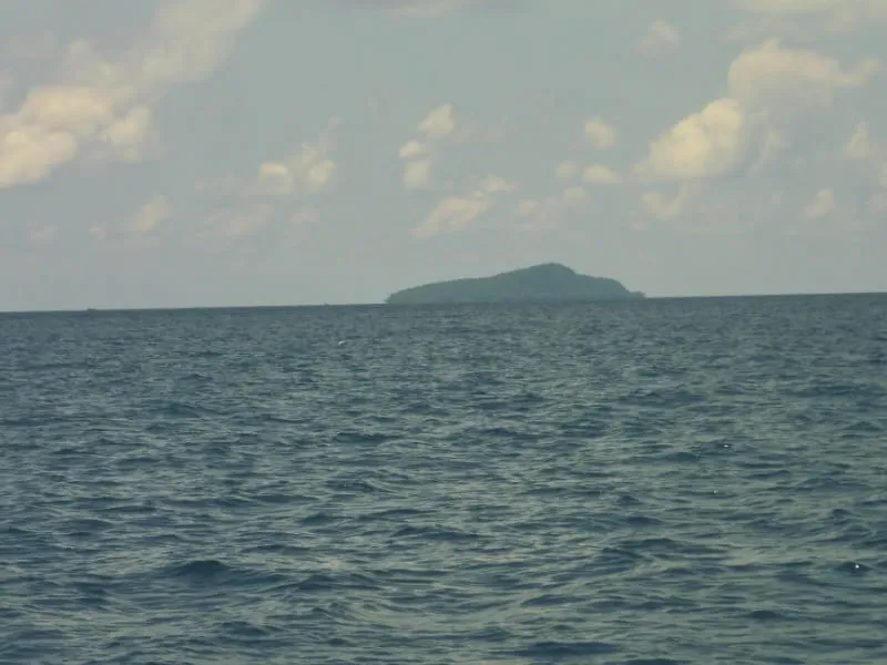 A wide shot of a calm blue ocean with a distant, green island under a partly cloudy sky.