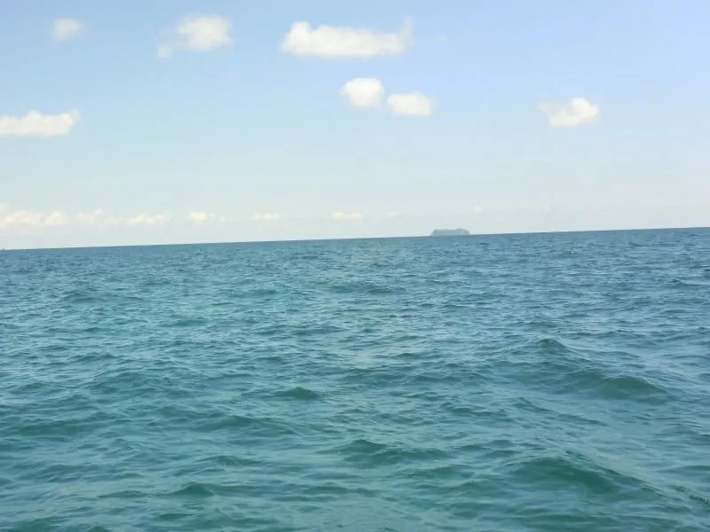 A wide shot of the blue-green ocean with gentle waves under a partly cloudy sky. A small island or landmass is visible on the distant horizon.