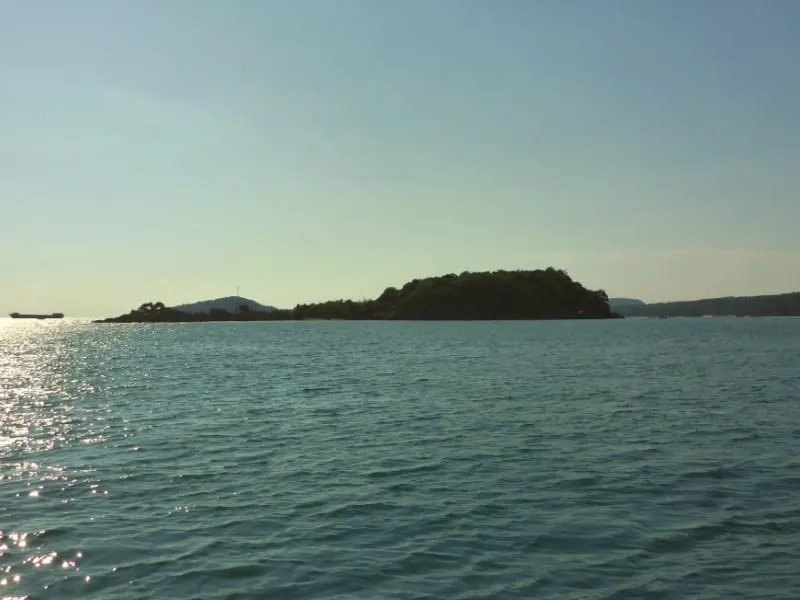 A wide shot of a calm blue sea with a lush green island in the distance under a clear sky. A ship is visible on the left horizon, with sunlight reflecting on the water.