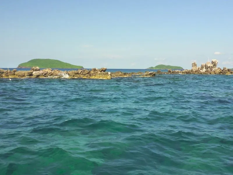 A clear blue-green ocean with rocky outcrops and lush green islands in the distance under a bright, clear sky.