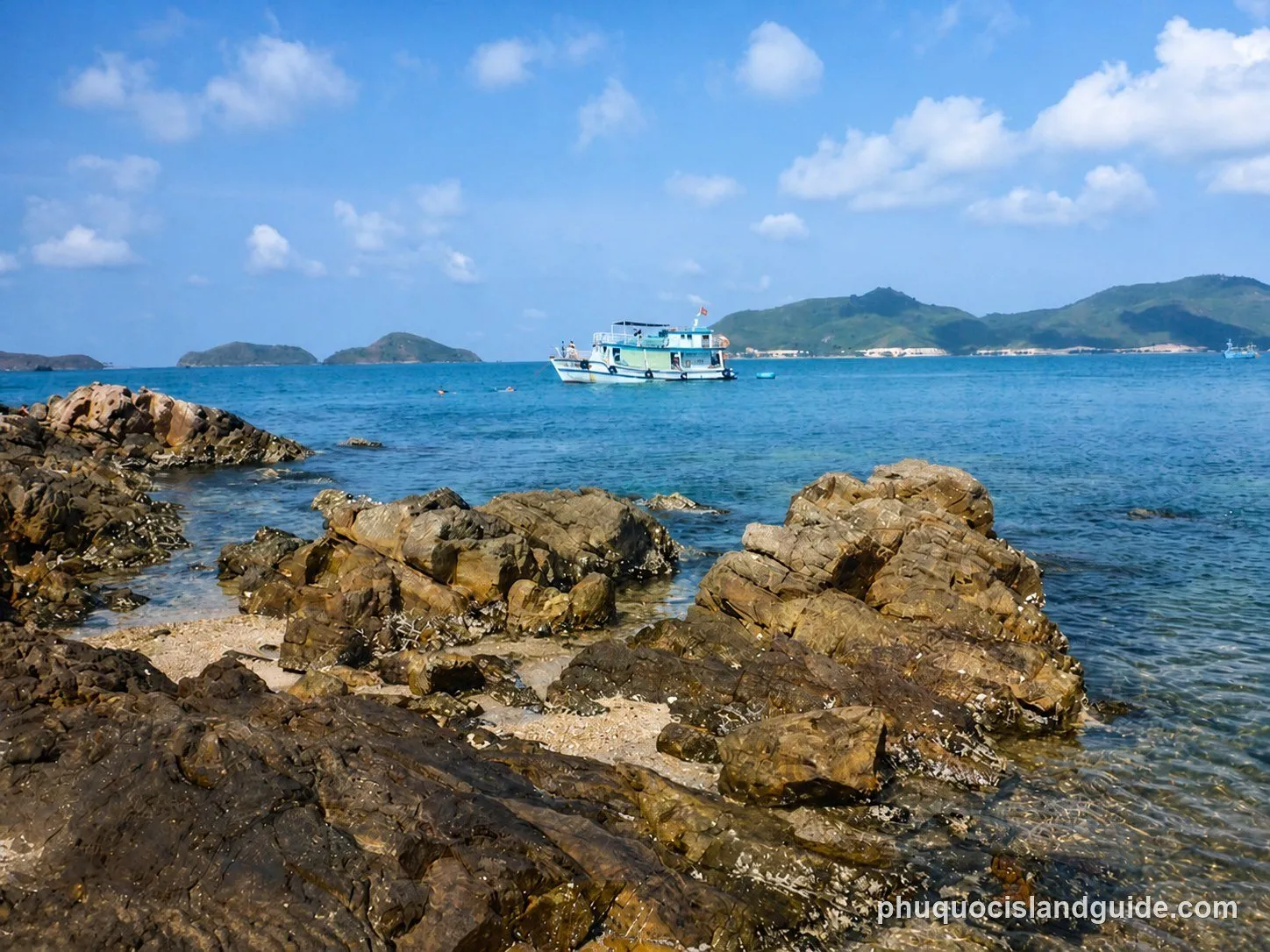 A scenic view of a rocky tropical coastline with clear blue water, a boat, and distant green islands under a bright blue sky with scattered clouds.