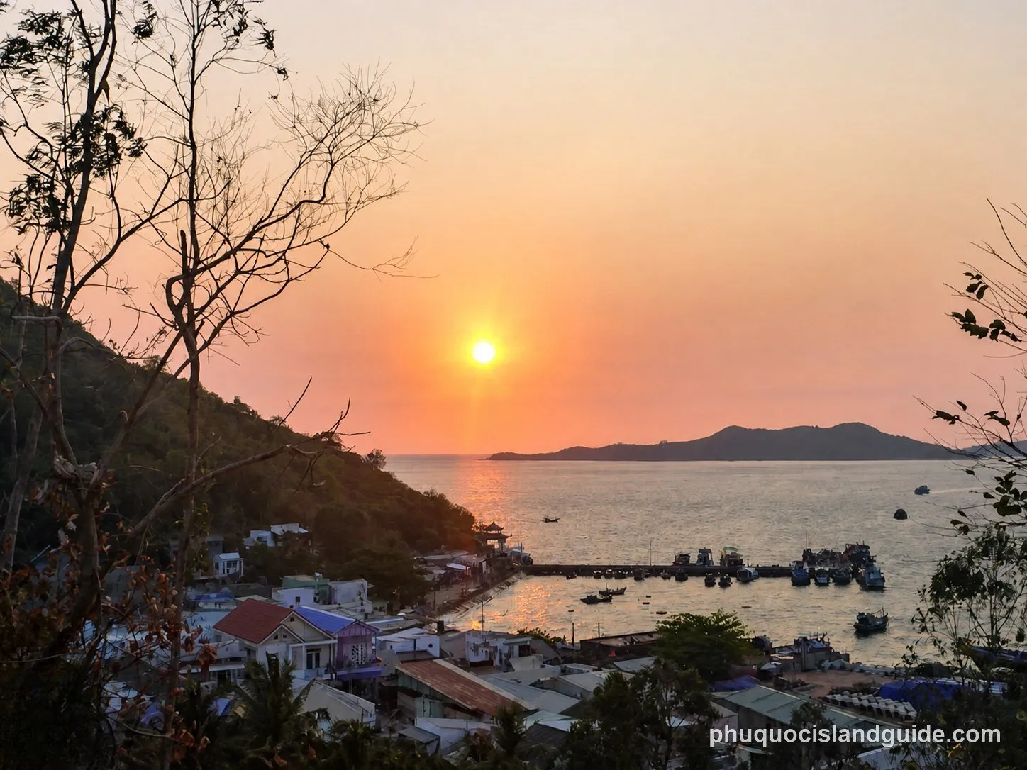 A scenic view of a sunset over a bay with a fishing village, a pier full of boats, and mountains in the background, seen from a hillside.