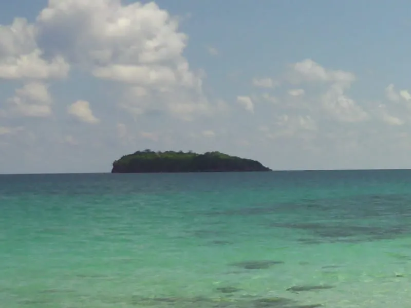 A small, lush green island sits in the middle of a clear, turquoise sea under a blue sky with scattered white clouds. The water is calm and transparent.