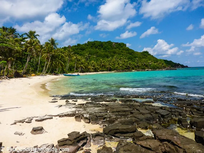A tropical beach with white sand, clear turquoise water, and palm trees. Dark rocks in the foreground, a small blue boat, and a green hill under a blue sky.