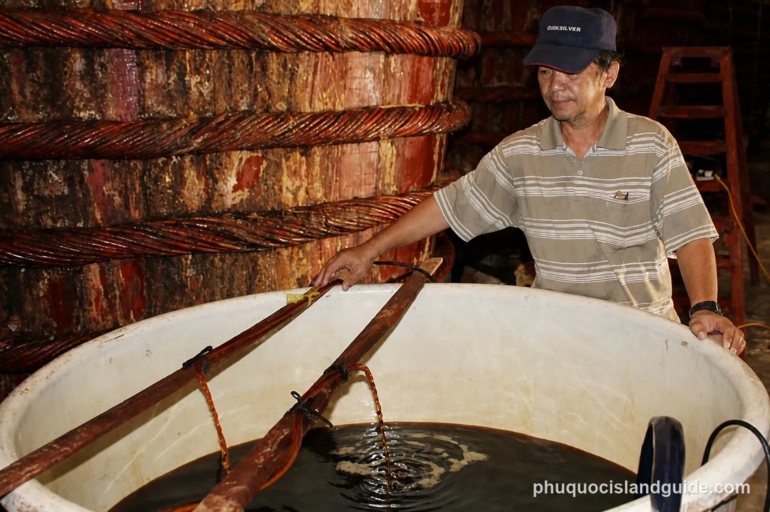 A man in a cap and striped shirt works with large vats, possibly for fish sauce production, with large wooden barrels in the background.