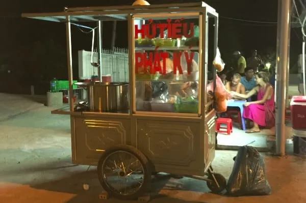 A street food cart with “HỦ TIẾU PHÁT KÝ” text, illuminated at night, with people dining at a small table in the background.