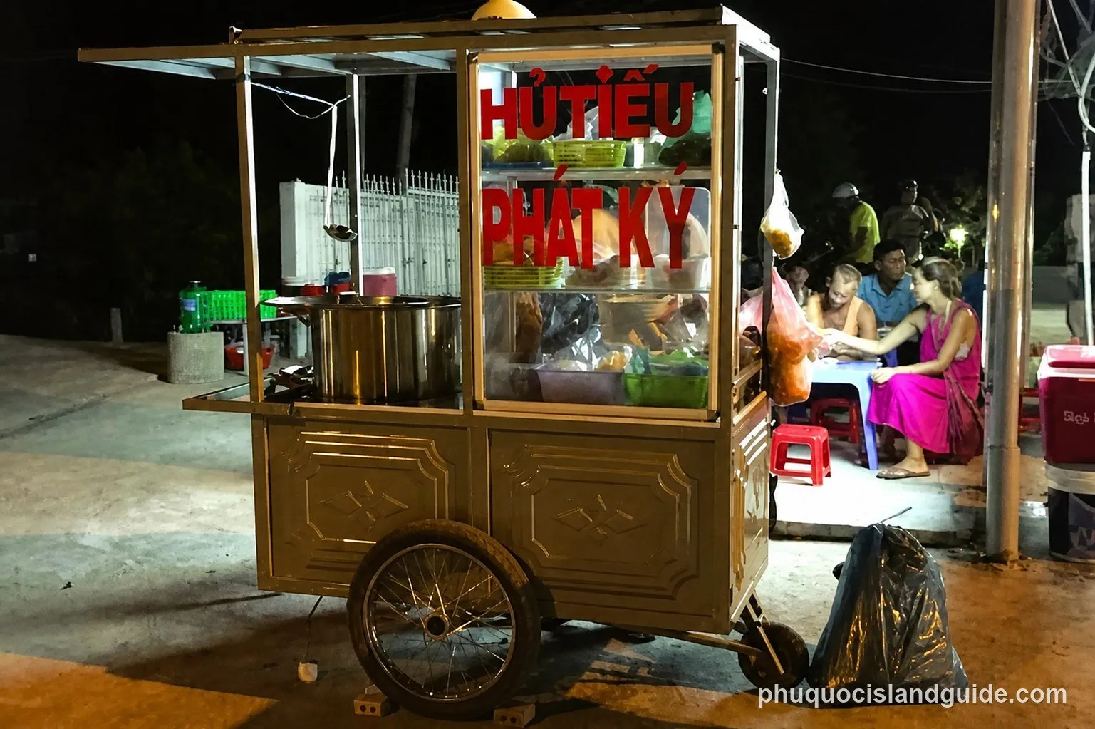 A street food cart with “HỦ TIẾU PHÁT KÝ” text, illuminated at night, with people dining at a small table in the background.