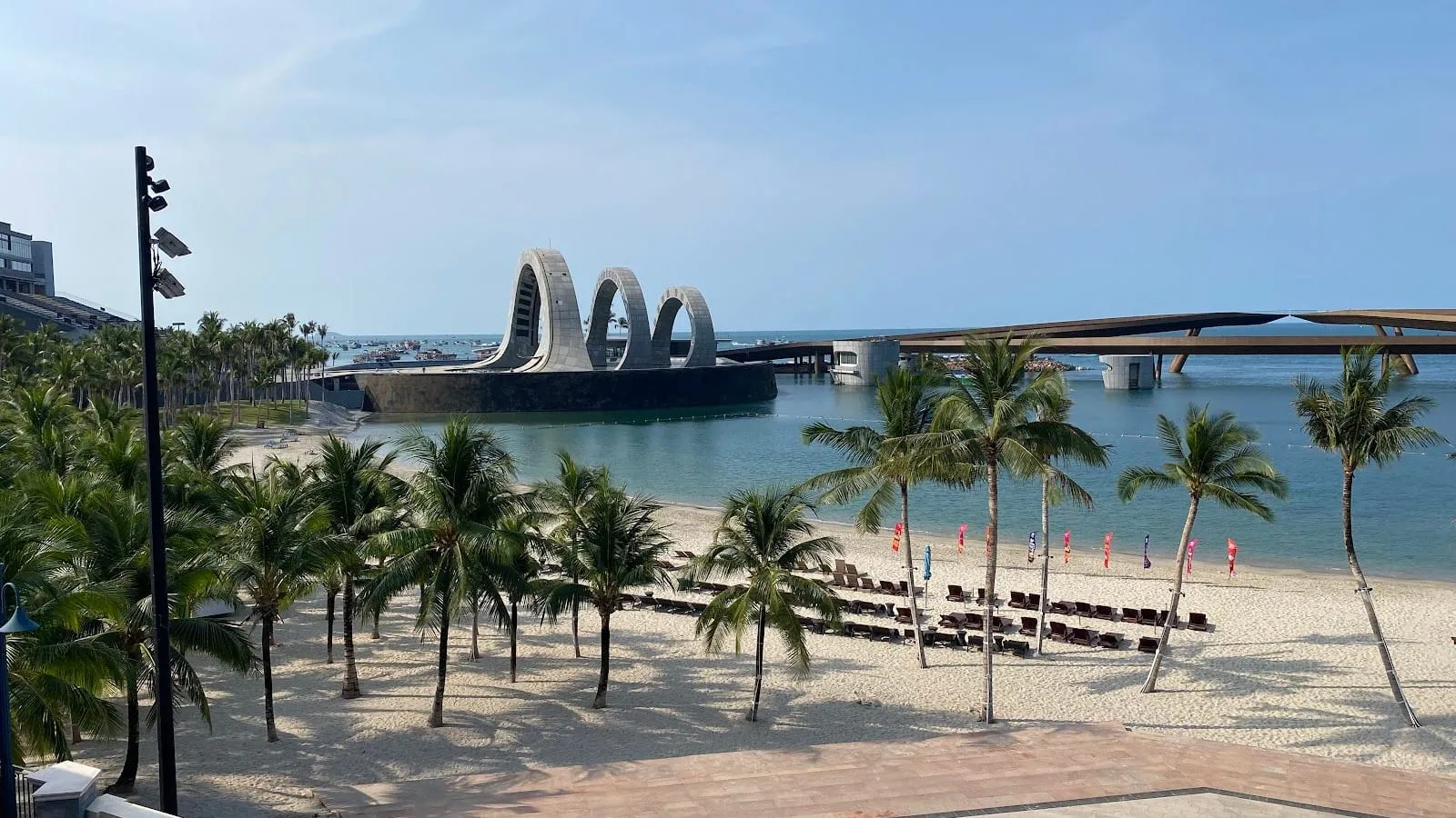Palm trees and sunbeds on the beach at Sunset Town, with the arches of the Kiss of the Sea stage and the Kiss Bridge behind, on a clear day in the dry season