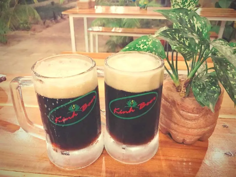 Two mugs of dark beer with foam on a wooden table, next to a potted green plant. The mugs have “Kinh Beer” written on them.