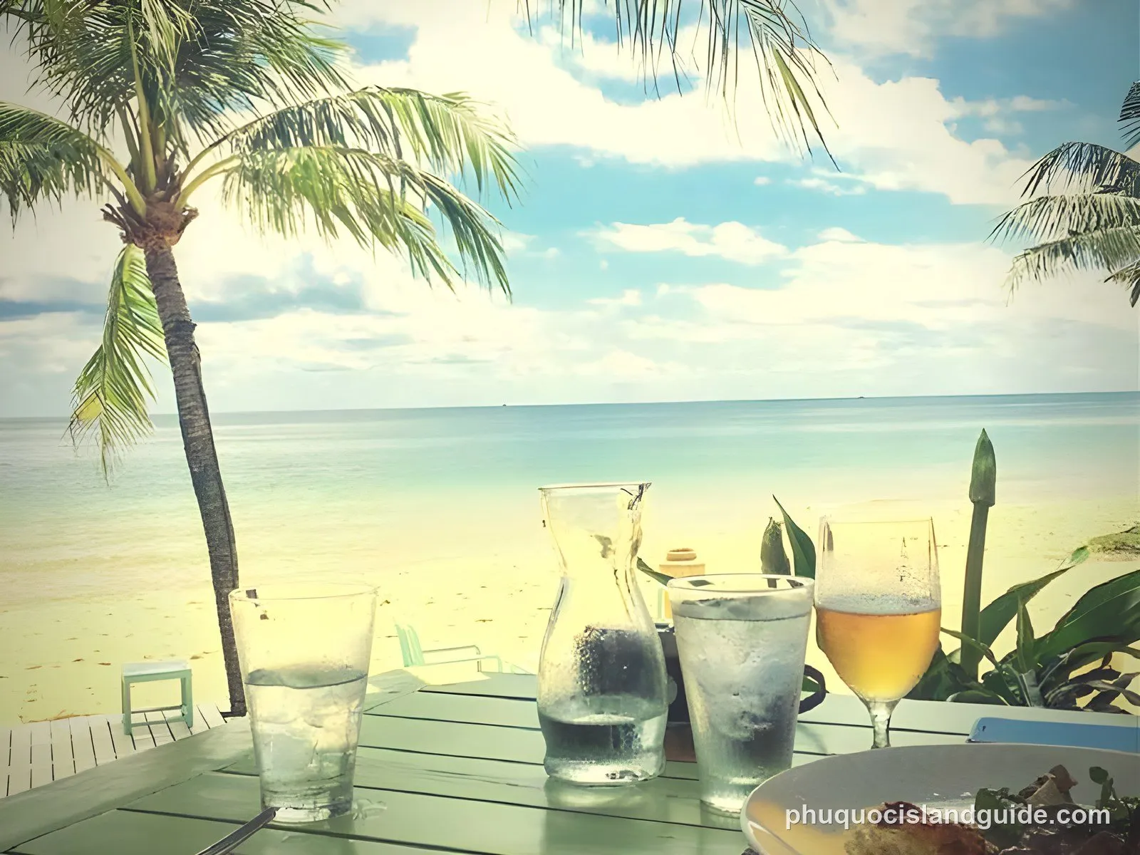A view from a table with drinks and a plate, looking out at a tropical beach with a palm tree and the ocean under a blue sky.