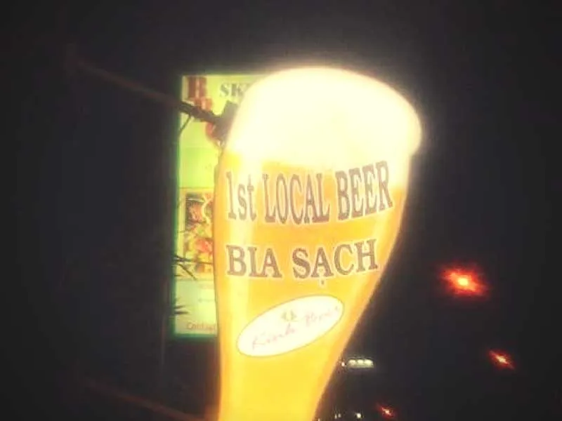 An illuminated outdoor sign shaped like a beer glass at night, advertising ‘1st LOCAL BEER BIA SẠCH’. The sign is bright yellow against a dark background.