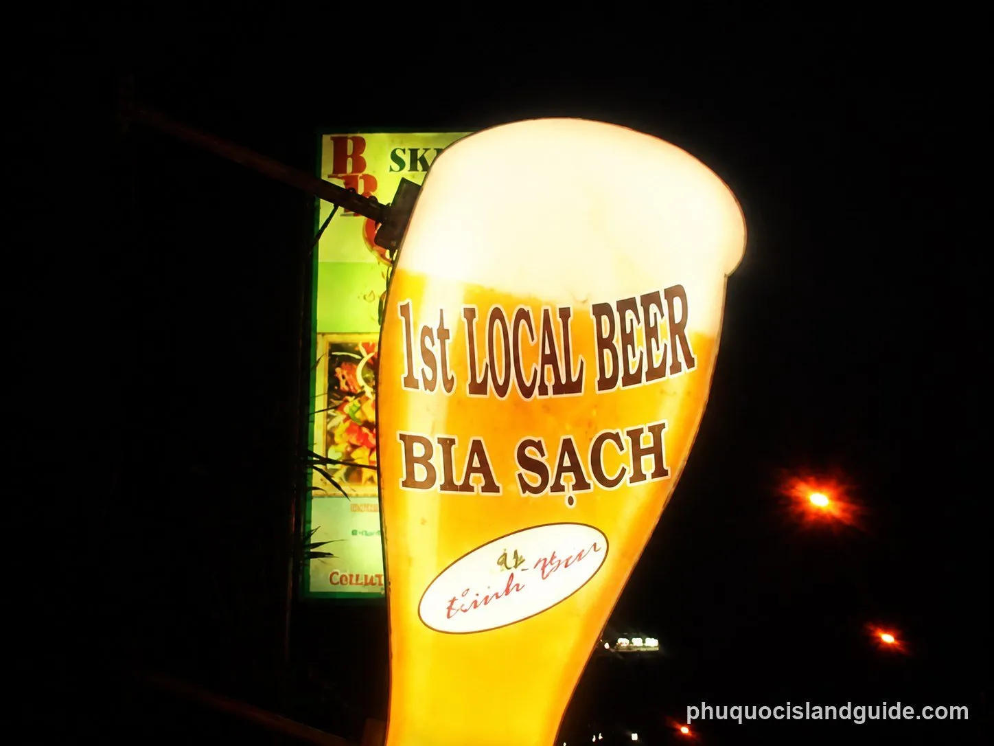 An illuminated outdoor sign shaped like a beer glass at night, advertising ‘1st LOCAL BEER BIA SẠCH’. The sign is bright yellow against a dark background.