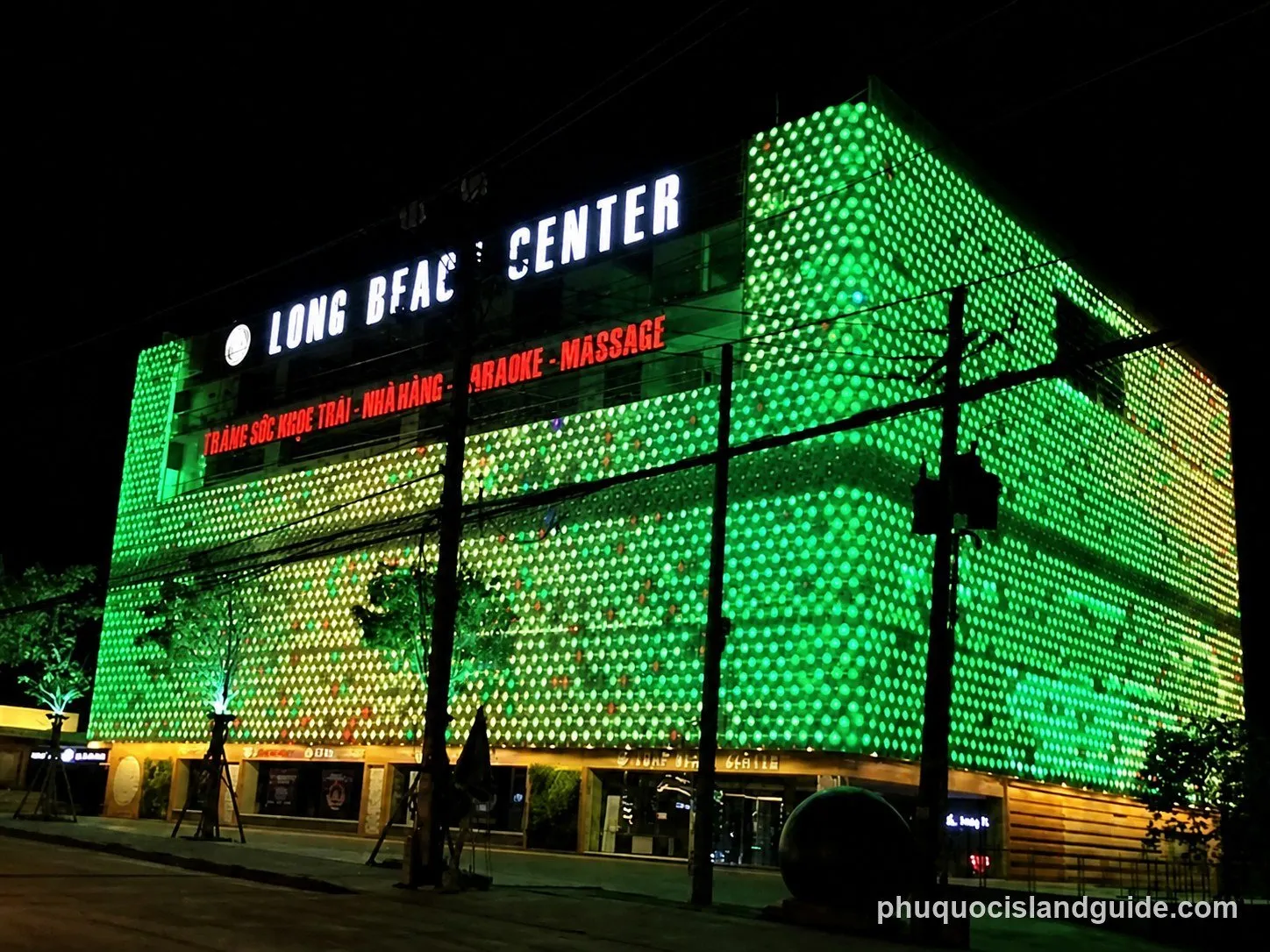 A large building illuminated with bright green and yellow lights at night. Signs read ‘LONG BFAC CENTER’ and list services like restaurant, karaoke, and massage.