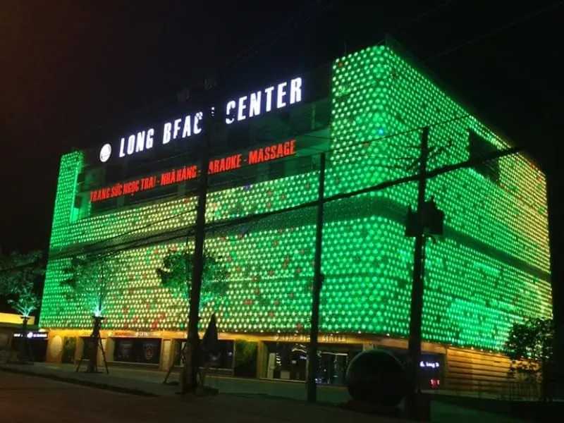 A large building illuminated with bright green and yellow lights at night. Signs read ‘LONG BFAC CENTER’ and list services like restaurant, karaoke, and massage.