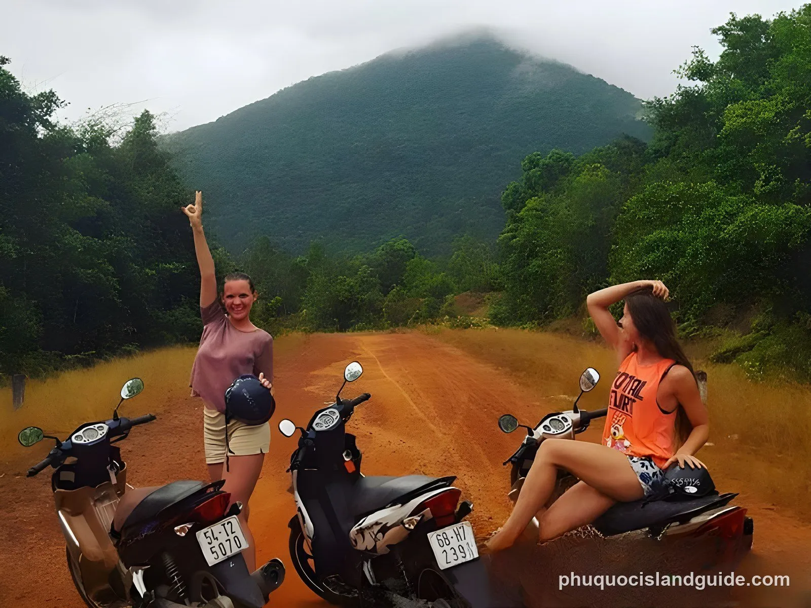 Two young women with three motorbikes on a dirt road, surrounded by lush green mountains and trees. One woman stands, making a peace sign; the other sits on a bike.