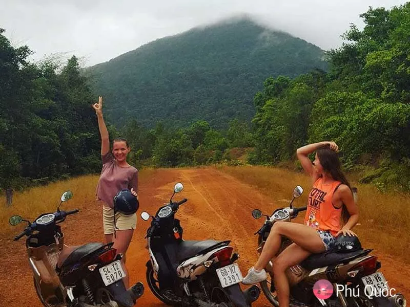Two young women with three motorbikes on a dirt road, surrounded by lush green mountains and trees. One woman stands, making a peace sign; the other sits on a bike.