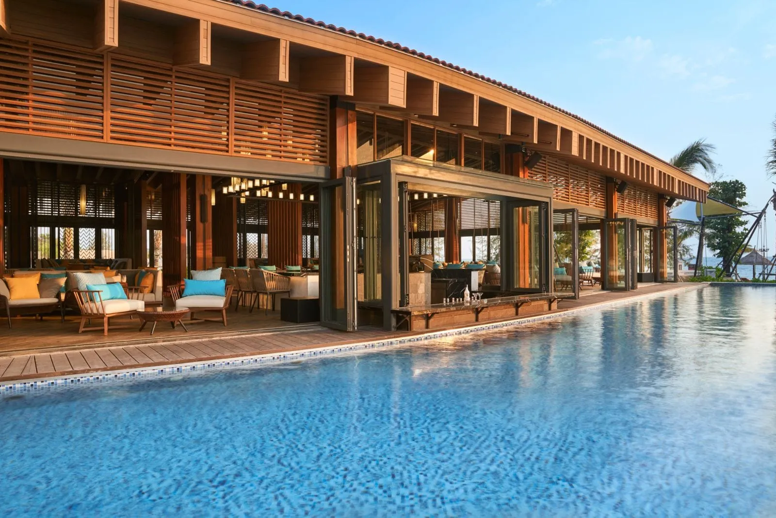 Poolside timber-slat pavilion with lounge seating opening onto a long swimming pool, with the sea beyond