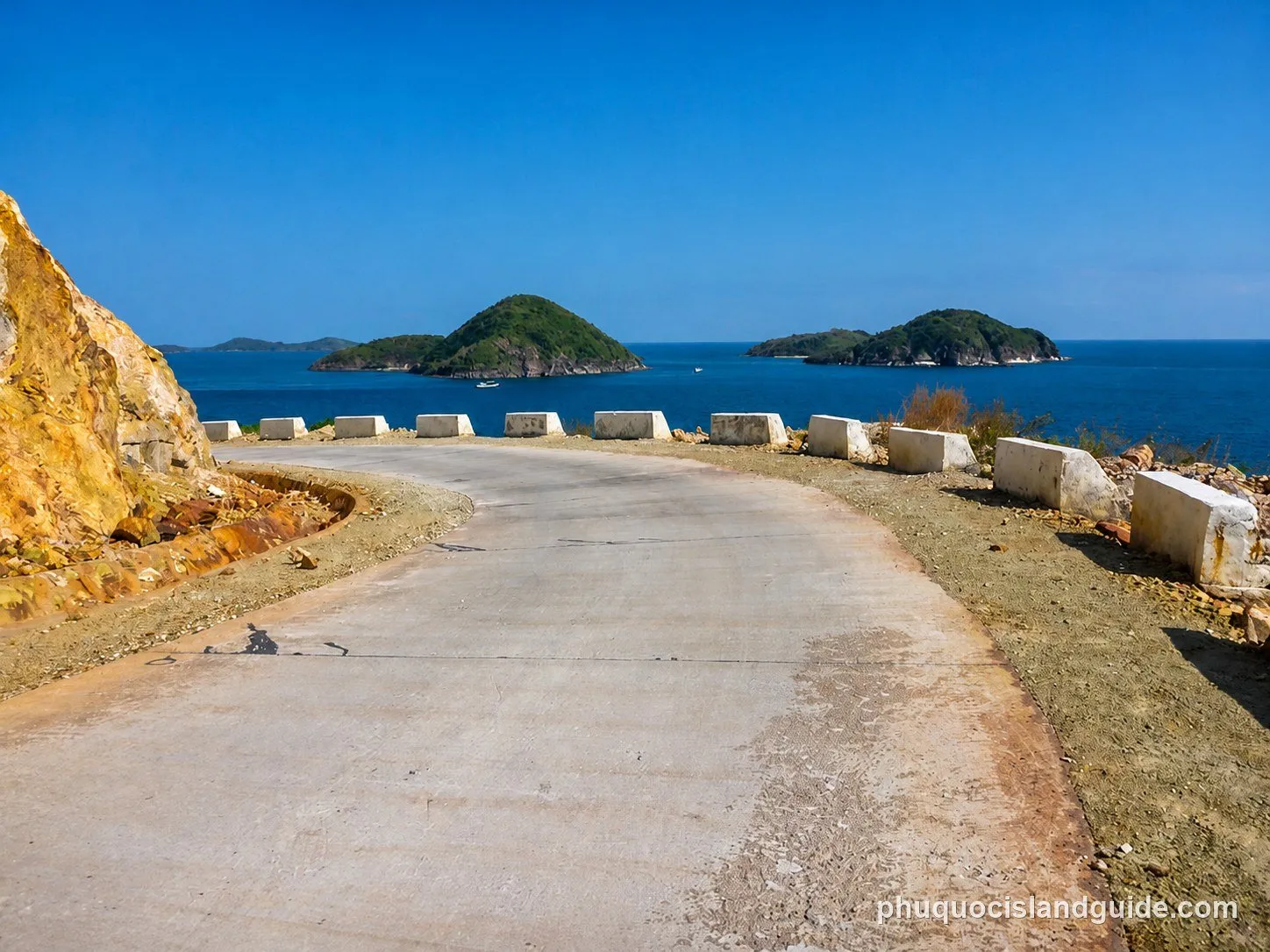 A winding coastal road with concrete barriers, overlooking green islands in a blue sea under a clear sky.