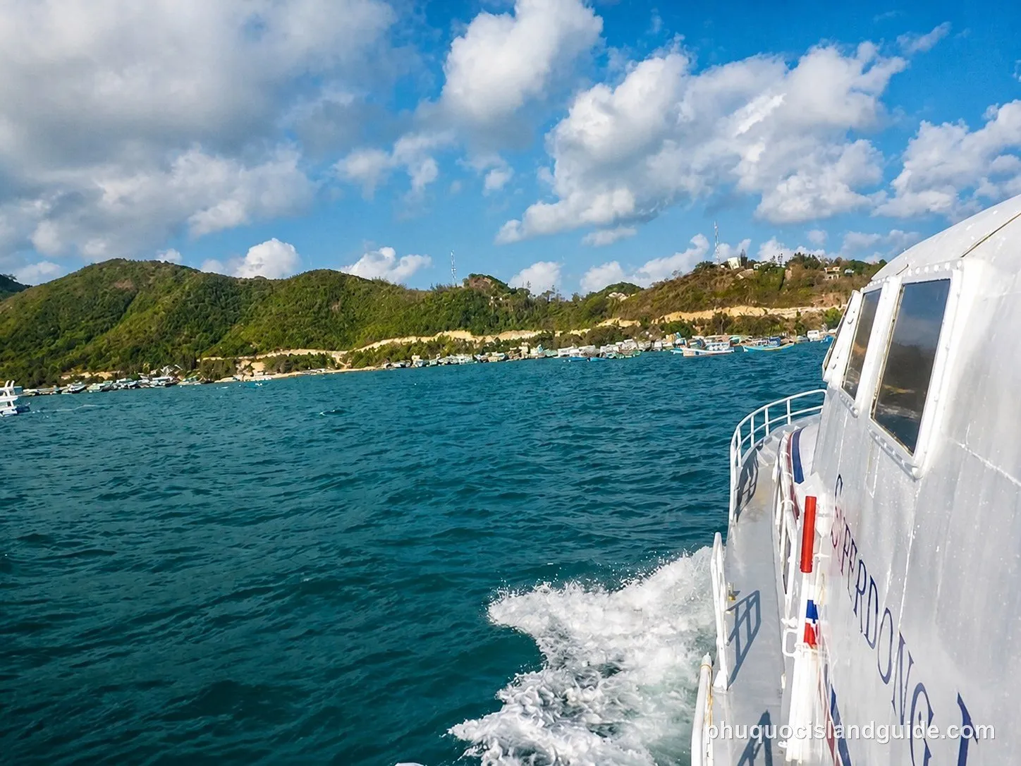 View from a boat moving through blue water, with a green, hilly coastline and a village in the background under a cloudy sky.