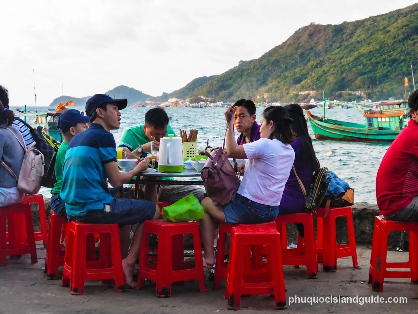 A group of people eating at an outdoor restaurant by the sea, sitting on red plastic stools. Fishing boats are visible in the water, with green mountains in the background.