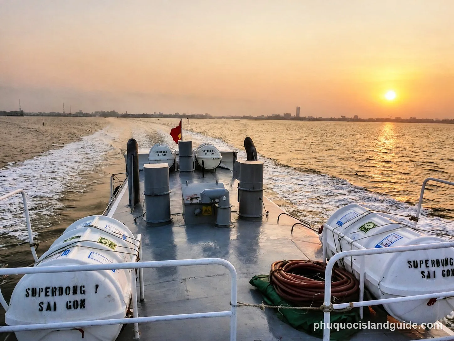 View from the stern of a boat at sunset, showing the wake, distant city skyline, and life rafts on deck. A Vietnamese flag flies at the back.