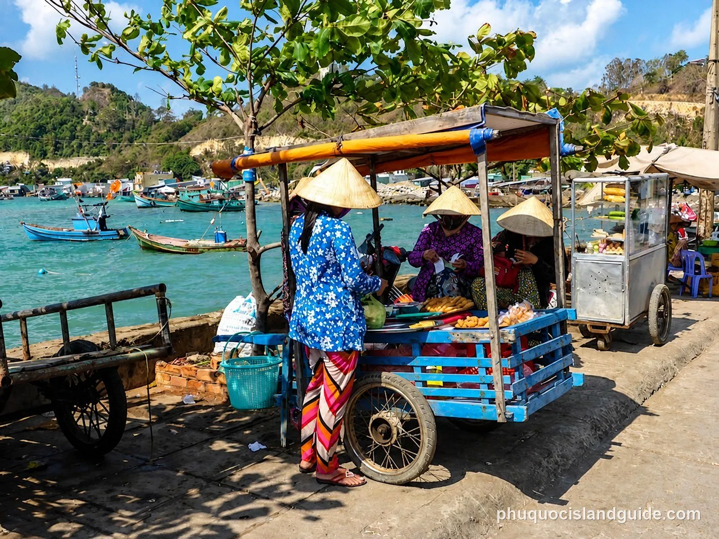 Three women in conical hats sell food from carts by the sea, with fishing boats and a green hillside in the background under a clear sky.