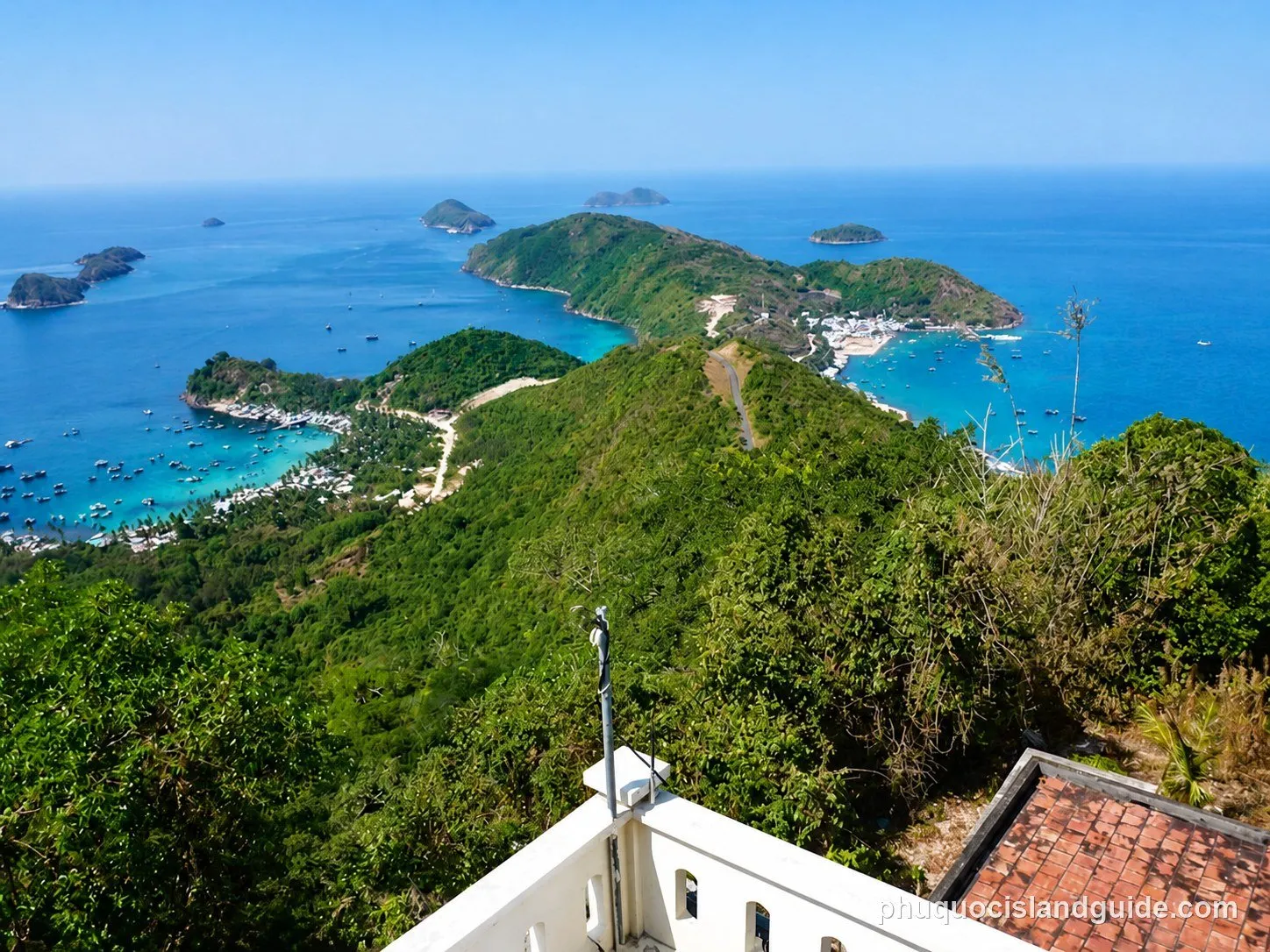 Panoramic view of a tropical bay with numerous small islands, lush green hills, a coastal village, and many boats anchored in the turquoise water.