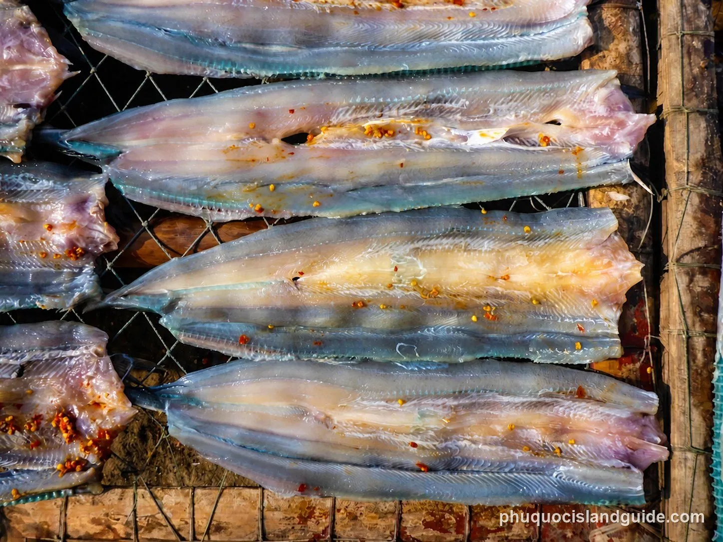 Close-up of several flattened, dried fish fillets, possibly seasoned with chili flakes, laid out on a mesh rack for sun-drying.