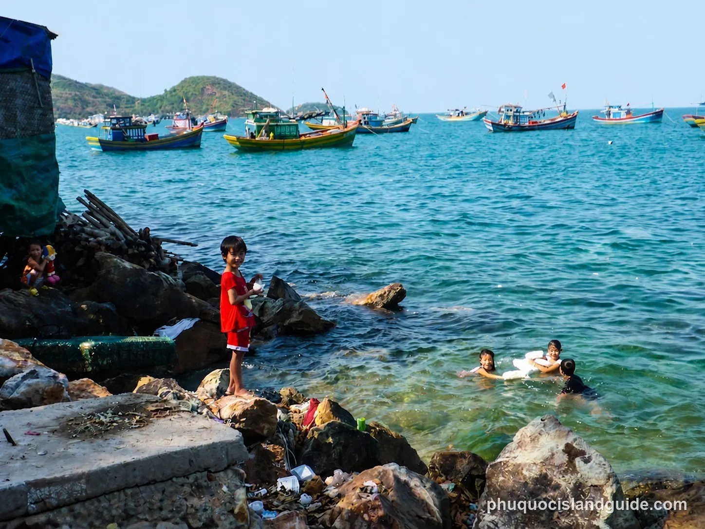 Children playing in the sea and on a rocky shore with fishing boats anchored in the background under a clear sky. Some debris is visible on the shore.