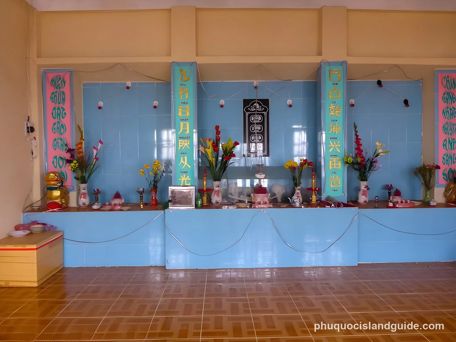 An indoor altar or shrine with blue tiled walls, decorated with vases of colorful flowers, offerings, and banners with Vietnamese text. A wooden box is on the left.