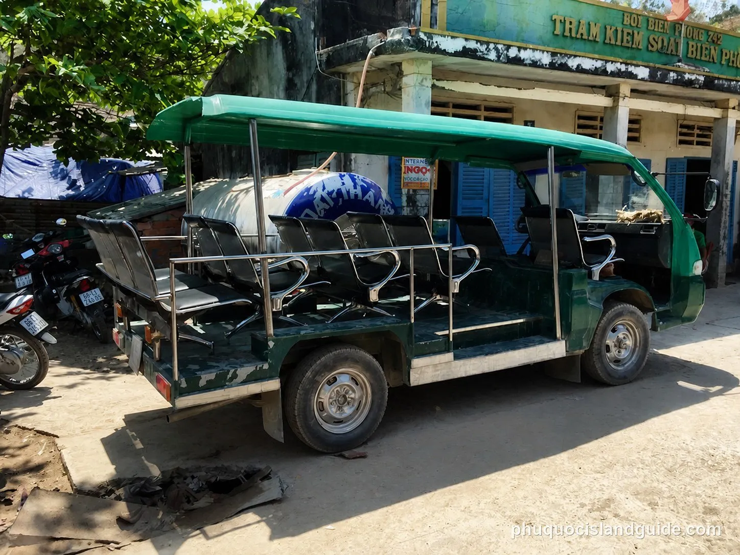 A green open-sided shuttle bus with multiple rows of seats is parked on a dirt road next to a building with Vietnamese text.