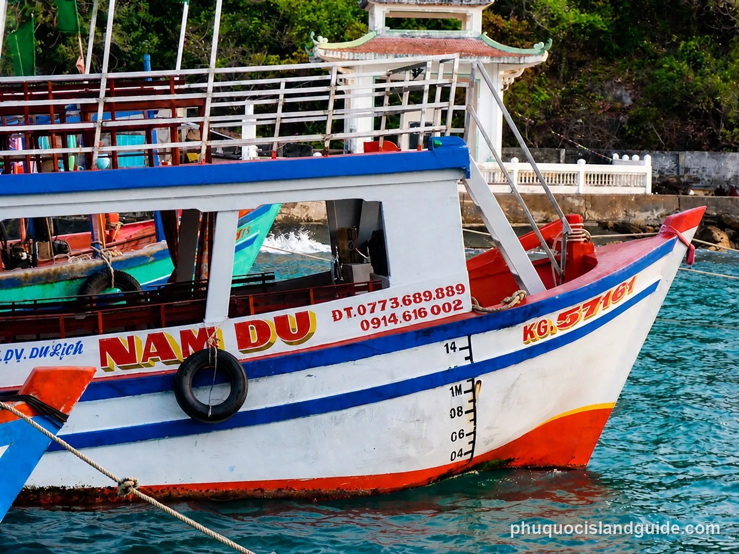 A close-up of the bow of a white, red, and blue boat named “NAM DU” docked in clear blue water, with another boat and a traditional white structure in the background.