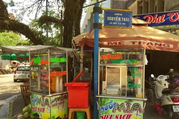 A street scene in Vietnam with two sugarcane juice stalls, a street sign for Nguyen Trai, parked vehicles, and lush green trees.