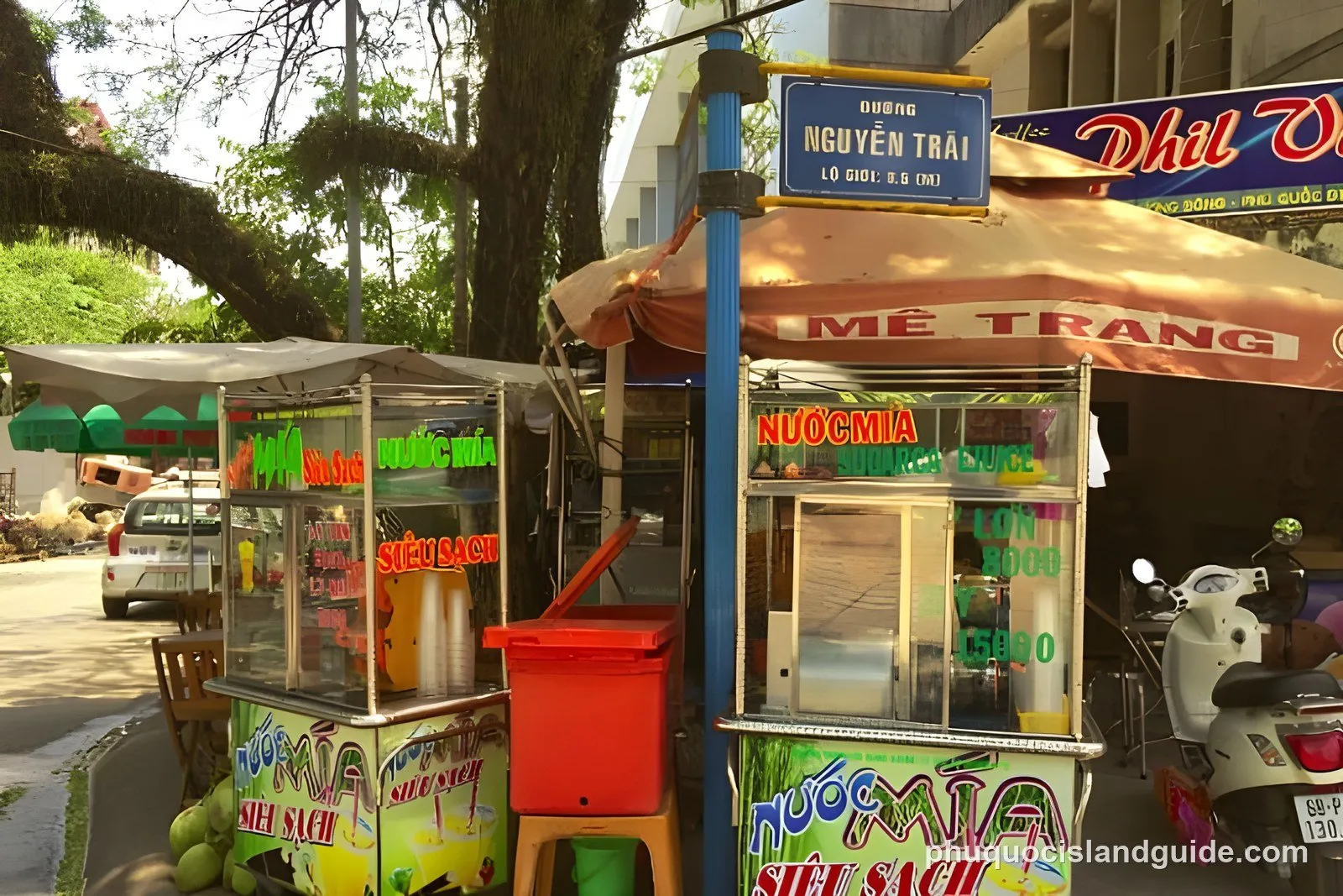 A street scene in Vietnam with two sugarcane juice stalls, a street sign for Nguyen Trai, parked vehicles, and lush green trees.