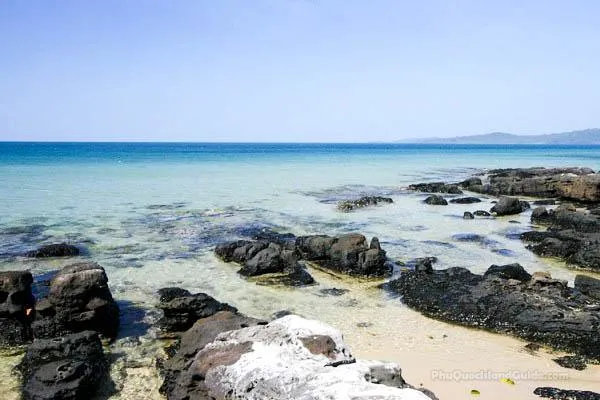 A clear blue sky over a calm, clear blue-green sea with dark rocks scattered along a sandy shore. Distant land is visible on the horizon.