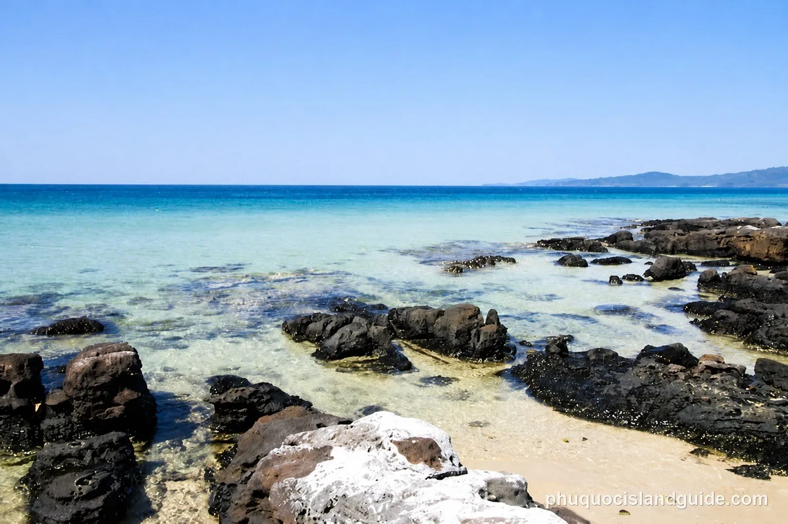 A clear blue sky over a calm, clear blue-green sea with dark rocks scattered along a sandy shore. Distant land is visible on the horizon.