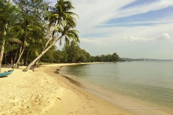 A tropical beach with golden sand, leaning palm trees, and lush green vegetation along the calm, clear water under a partly cloudy sky. A blue boat rests on the sand.