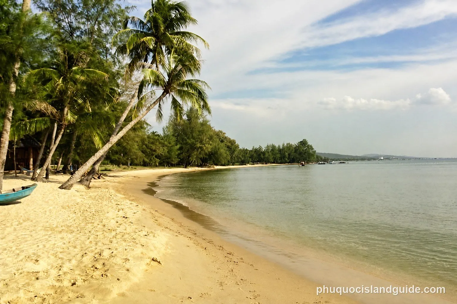A tropical beach with golden sand, leaning palm trees, and lush green vegetation along the calm, clear water under a partly cloudy sky. A blue boat rests on the sand.