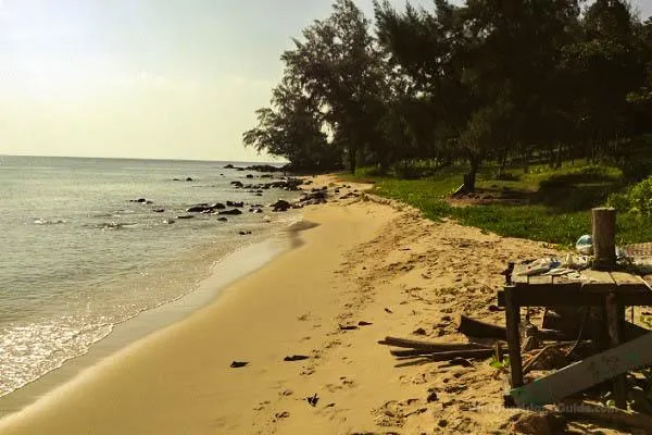 A sandy beach with gentle waves, scattered rocks in the water, and a line of green trees along the shore. A wooden structure is visible on the right.
