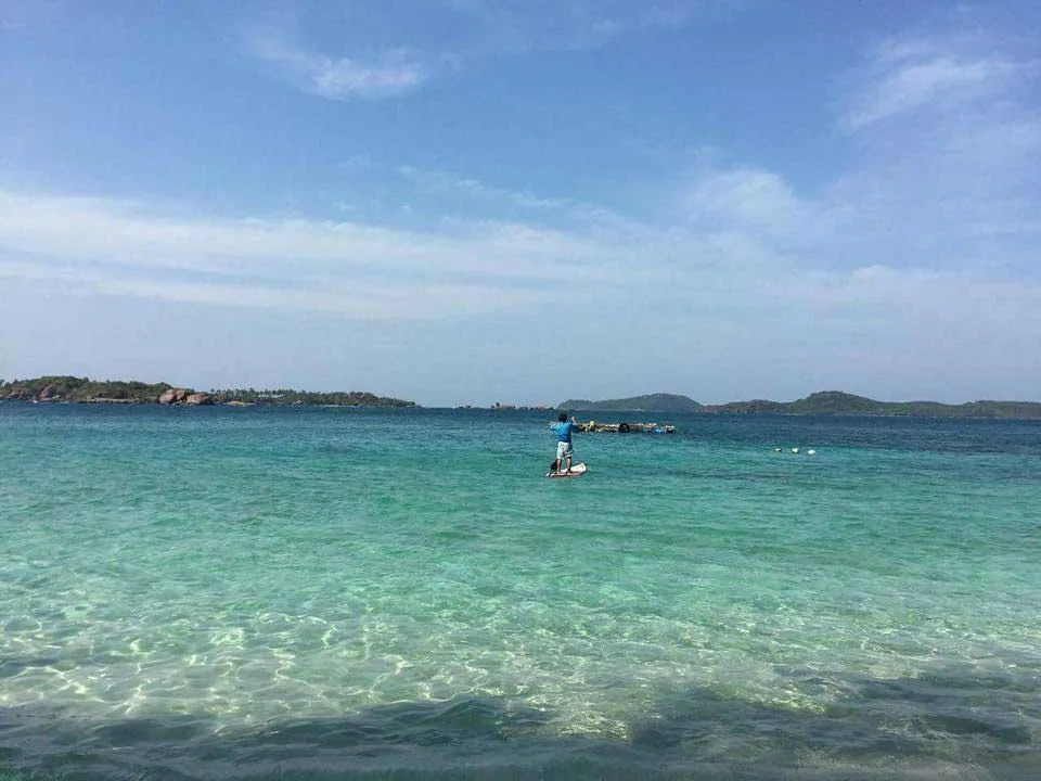 A person paddleboarding in clear turquoise ocean water with distant green islands under a blue sky.