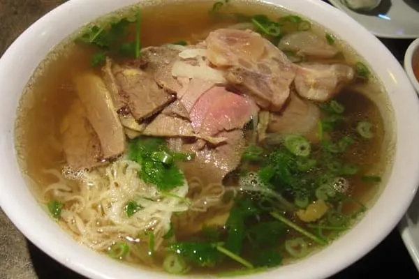 A close-up shot of a bowl of Vietnamese pho with various cuts of beef, rice noodles, and fresh green herbs in a clear broth.