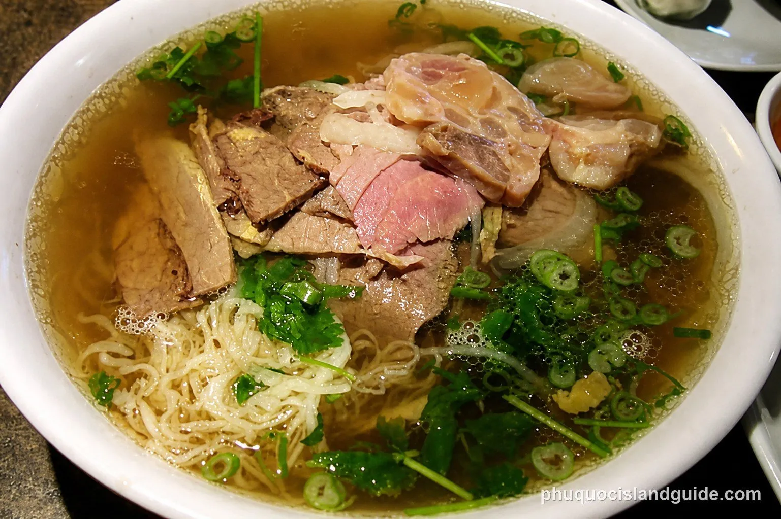 A close-up shot of a bowl of Vietnamese pho with various cuts of beef, rice noodles, and fresh green herbs in a clear broth.