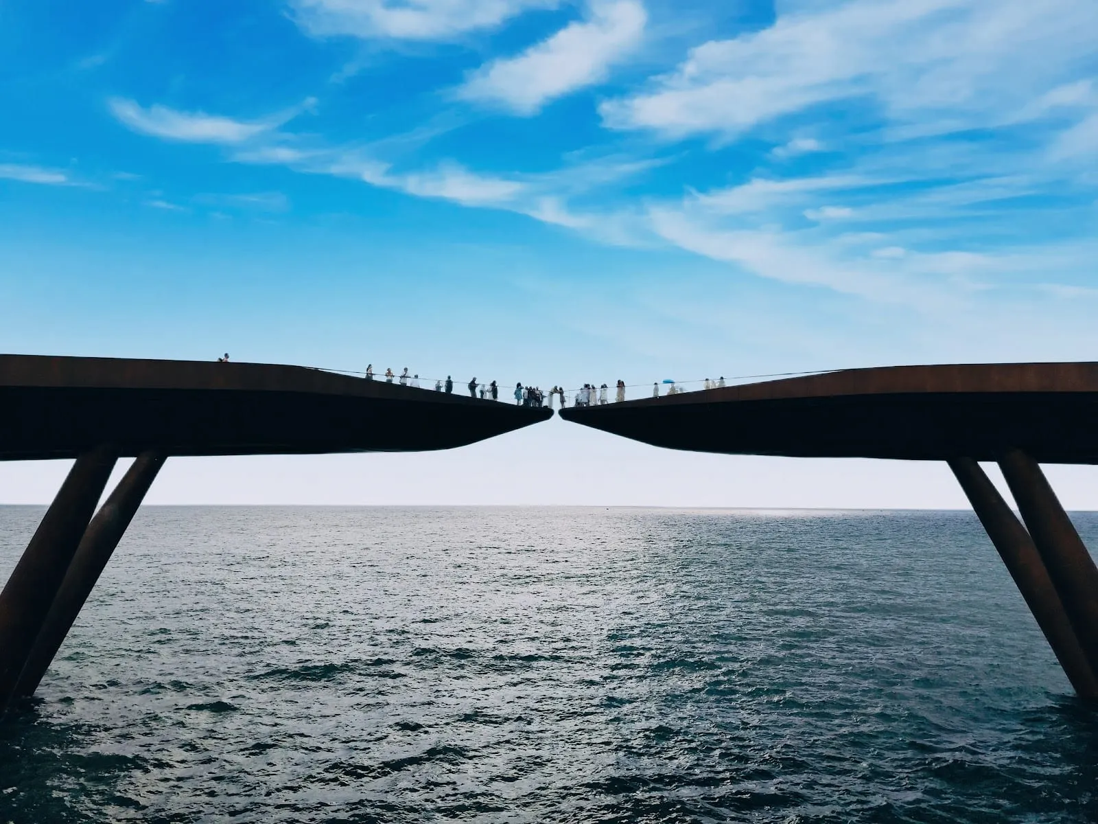 The two spans of the Kiss Bridge reaching towards each other over the sea at Sunset Town, under a blue sky
