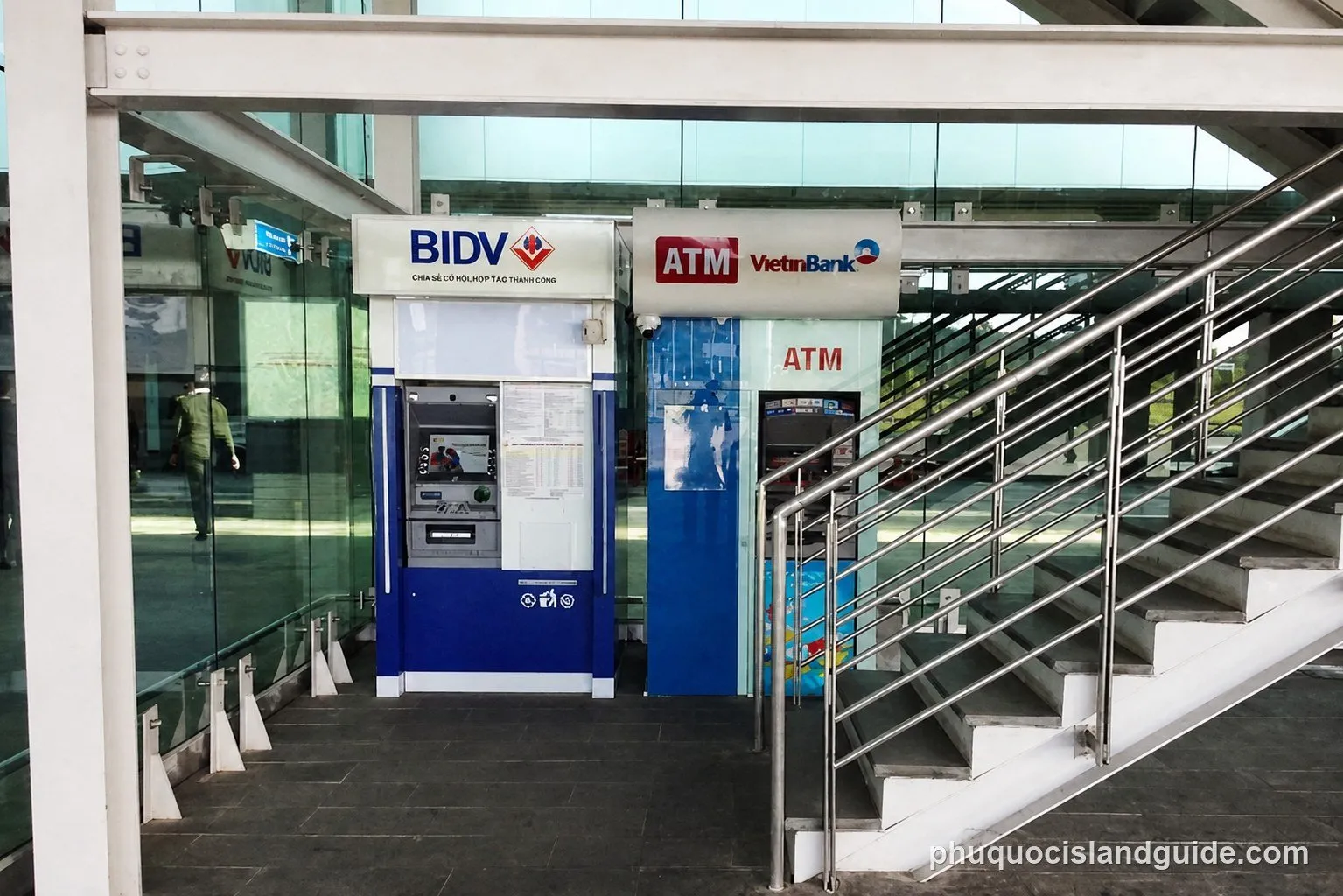 Two bank ATM machines beside a metal staircase inside the airport terminal, with a person walking past in the background.