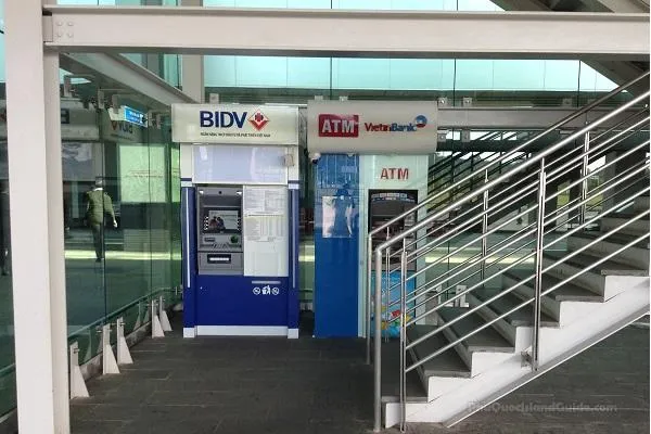 Two bank ATM machines beside a metal staircase inside the airport terminal, with a person walking past in the background.